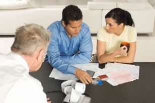 Three people sit at a table, discussing paperwork. One man in a blue shirt points at documents, while a woman holding a mug listens. Another man sits with his back to the camera. A calculator and papers are on the table.