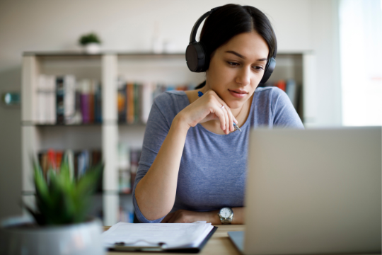 A woman wearing headphones sits at a desk, looking at a laptop screen. She has a notepad and papers in front of her, and bookshelves are visible in the background.