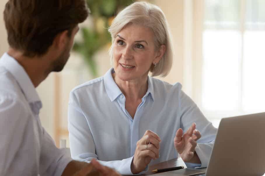 An older woman with short gray hair talks to a younger man while sitting at a table with a laptop. She gestures with her hands and appears engaged in conversation in a bright, indoor setting.