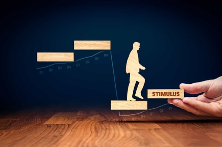 A hand holds a wooden block labeled STIMULUS, helping a wooden figure of a person step upward onto higher blocks, symbolizing economic support and growth.