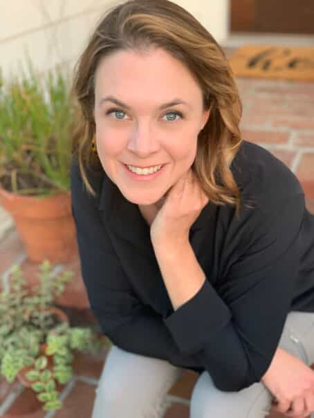 A woman with light brown hair and a black shirt smiles up at the camera while sitting on brick steps next to potted plants.