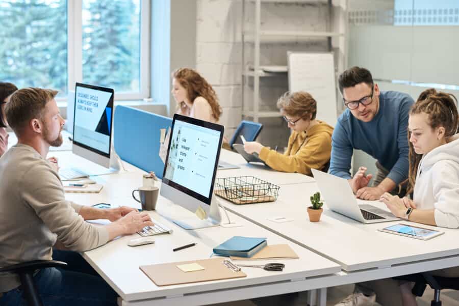 A group of people work together at a large desk in a modern, bright office. Some use computers and laptops, while others write or discuss tasks. Office supplies and potted plants are on the desks.