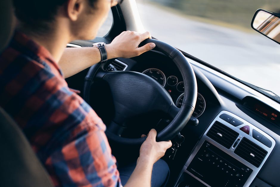 A person wearing a red and blue plaid shirt is driving a car, holding the steering wheel with both hands. The dashboard and speedometer are visible, and the road can be seen through the windshield.