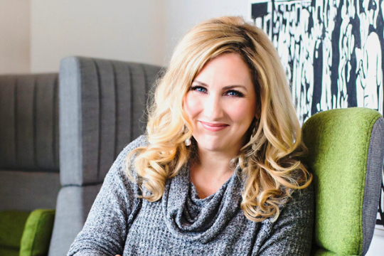 A woman with long blonde curly hair and a grey sweater smiles while sitting on a green chair. The background features modern gray furniture and black-and-white wall art.