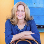A woman with long blonde hair, wearing a blue top, sits with her arms resting on a chair. She is smiling and posed in front of a yellow wall and a blue wooden door.