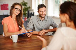 Three people sit around a wooden table in an office setting. One woman takes notes on a clipboard, a man sits beside her with a laptop, and another woman speaks to them with her back to the camera.
