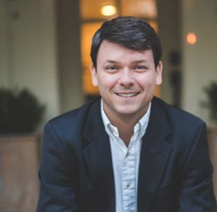 A man with short dark hair, wearing a white shirt and dark blazer, is smiling at the camera. He is sitting outdoors with a blurred background and warm lighting.
