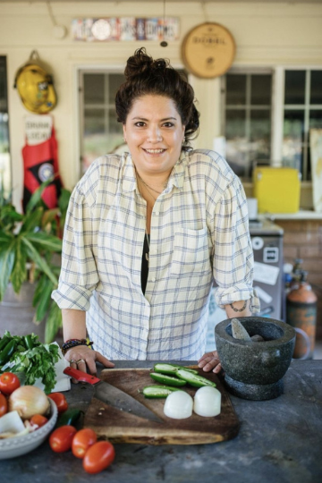 A woman in a plaid shirt smiles at the camera while standing in a kitchen. On the counter in front of her are fresh vegetables, a knife, and a mortar and pestle. The background shows plants and various kitchen items.