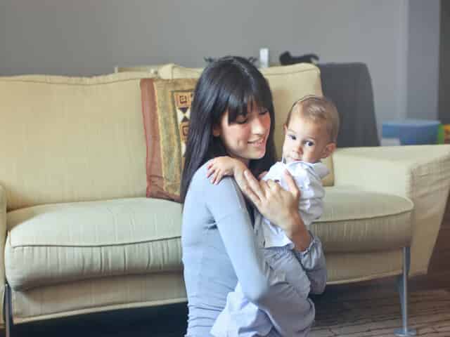A woman with long dark hair smiles while holding a baby dressed in light-colored clothes, sitting on the floor in front of a beige sofa inside a living room.