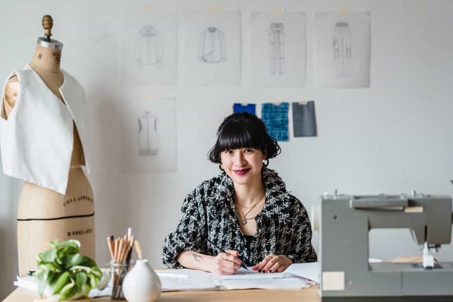 A smiling woman sits at a desk with design sketches, fabric swatches, a sewing machine, and a dress form in a fashion studio, working on sketches or notes.