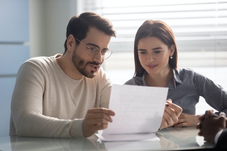 A man and a woman sit at a table, looking at a document together and discussing it. Both appear focused and engaged, with light coming in through a window behind them.