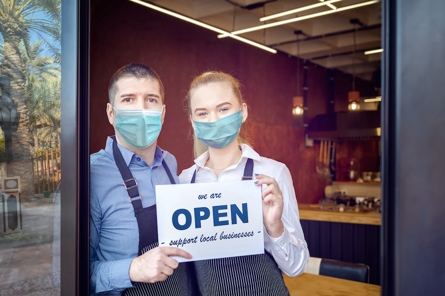 Two cafe workers wearing face masks stand in a doorway, holding a sign that reads We are OPEN support local business. The interior of the cafe and some palm trees outside are visible in the background.