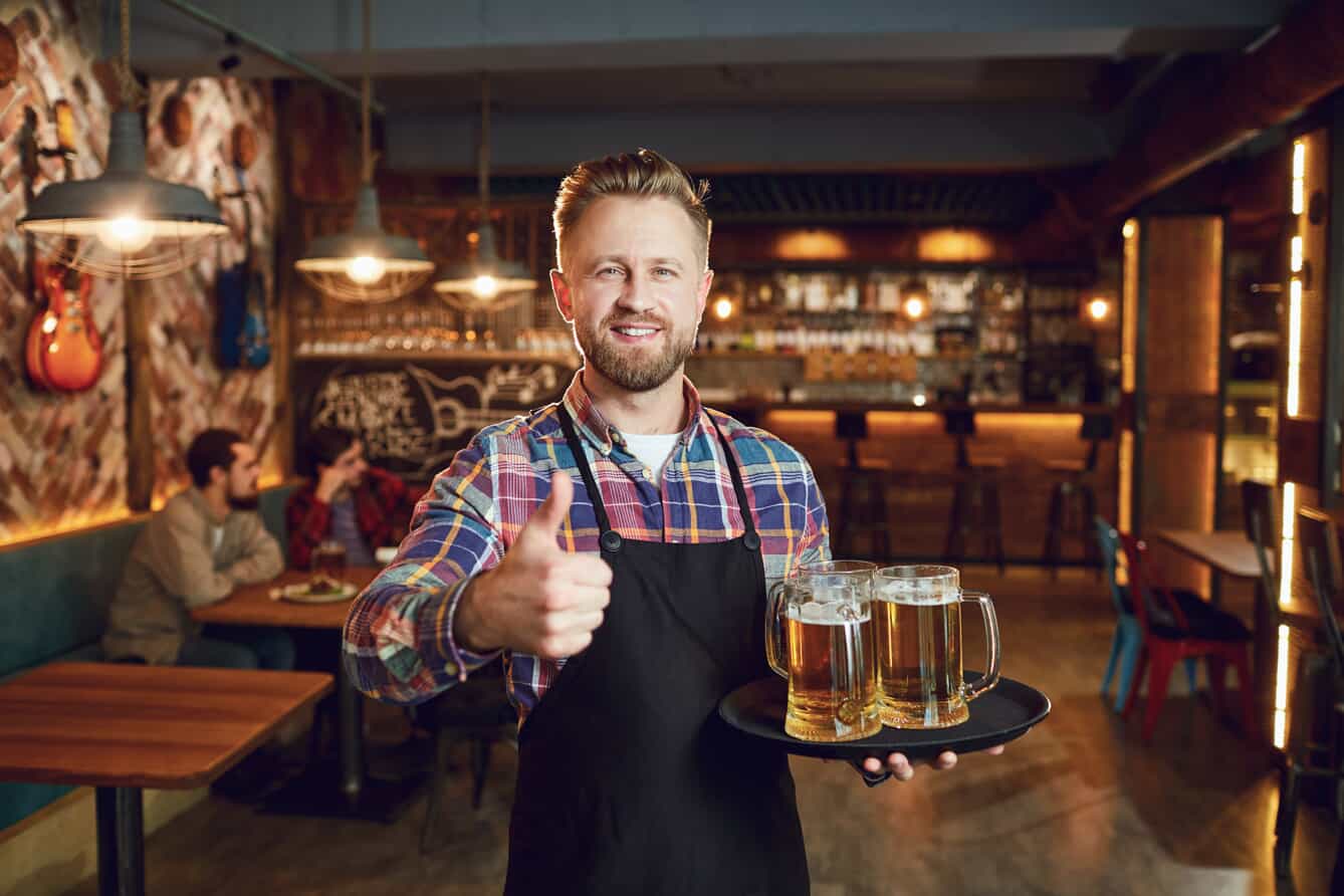 A smiling waiter wearing a plaid shirt and black apron holds a tray with four mugs of beer in one hand and gives a thumbs up with the other, standing in a cozy bar with people in the background.