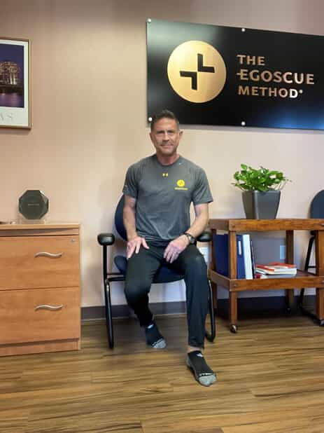 A man sits on a chair indoors, demonstrating an exercise move. He wears a gray T-shirt, black pants, and socks. Behind him is a sign reading The Egoscue Method and a small plant on a wooden table.