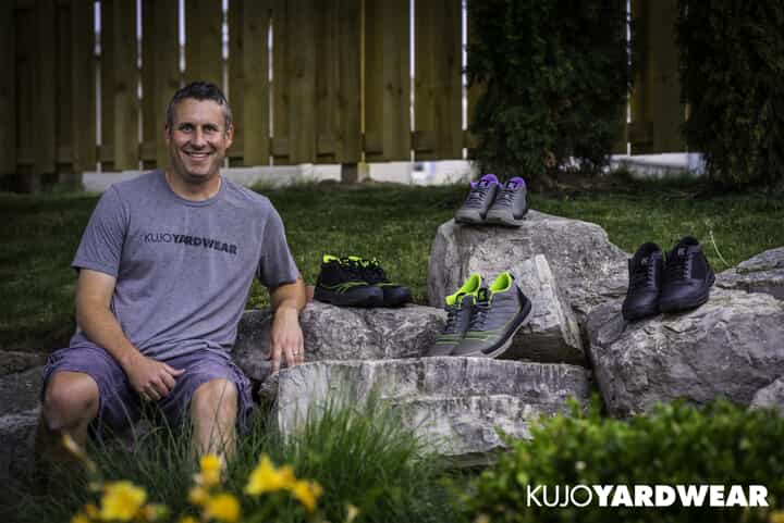 A man in a Kujo Yardwear shirt sits on rocks in a yard, smiling. Four pairs of yard shoes are displayed on the rocks around him. Green grass and yellow flowers are in the foreground. Kujo Yardwear logo is in the bottom right corner.