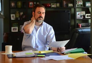 A man sits at a cluttered table, holding his neck with one hand and looking at a piece of paper in the other, appearing stressed or tired. There are folders, papers, and a coffee mug on the table.