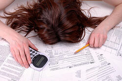 A person with long brown hair rests their head face down on a pile of tax forms, holding a pencil in one hand and a calculator in the other, appearing overwhelmed and exhausted.