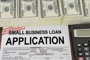 A small business loan application form marked approved is shown with stacks of $100 bills and part of a calculator visible on the side.