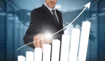 A man in a suit is touching a glowing bar on a rising bar graph with an upward arrow, symbolizing business growth and success. The background shows a blurred world map and office setting.