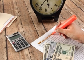 A person highlights text on a document beside a calculator, U.S. dollar bills, and a clock on a wooden table.