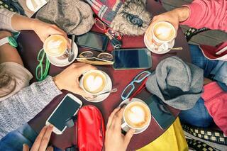 Four people sit around a table with lattes, each holding a cup. The table is scattered with smartphones, glasses, hats, and accessories. Seen from above, the scene is cozy and casual.