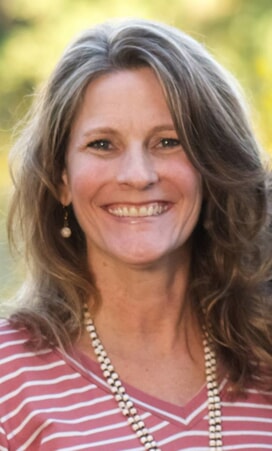 A woman with wavy, light brown hair smiles at the camera. She is wearing a pink and white striped top, a beaded necklace, and pearl earrings. The background is outdoors and softly blurred.