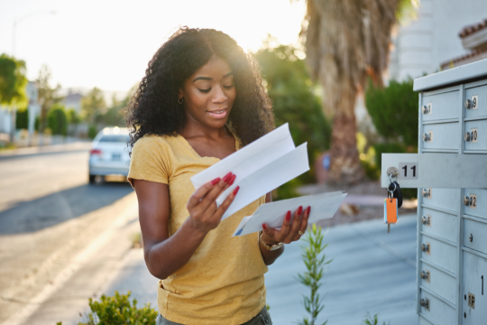 A woman stands outdoors by a row of mailboxes, smiling as she reads a letter. She wears a yellow shirt and holds several envelopes. The sun is shining, and palm trees and houses are visible in the background.