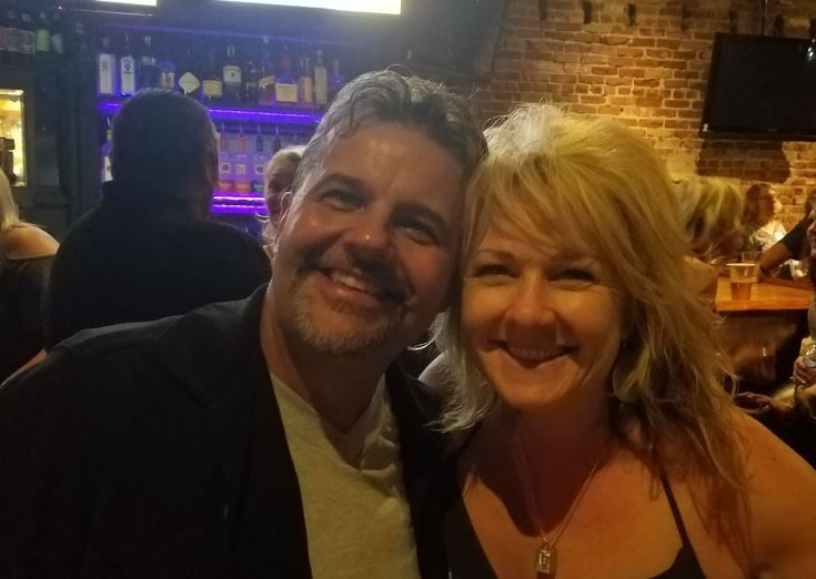A smiling man and woman pose closely together in a dimly lit bar with exposed brick walls and bottles on shelves in the background. Other people sit at tables, and the atmosphere appears casual and cheerful.