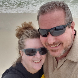 A smiling couple wearing sunglasses takes a selfie on a beach, with waves and sand visible in the background. The woman has curly hair in a bun, and the man has short hair and a trimmed beard.