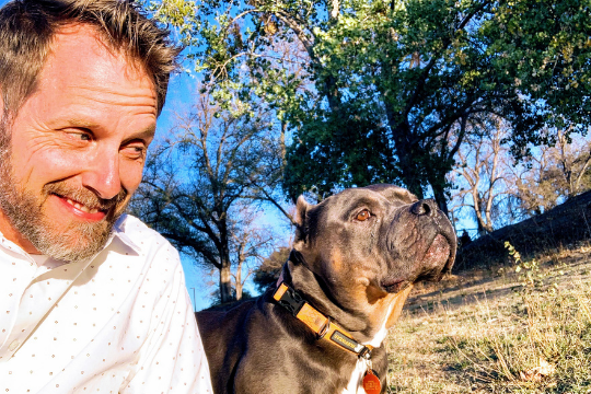A man in a white shirt smiles while sitting outdoors next to a brown and gray dog wearing a collar. Trees and sunlight fill the background, creating a cheerful and relaxed atmosphere.