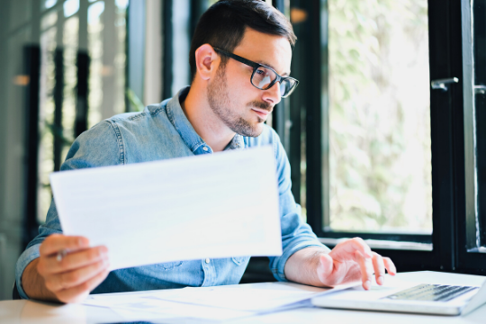 A man wearing glasses and a denim shirt sits at a desk by a window, holding a sheet of paper in one hand and typing on a laptop with the other, appearing focused on his work.