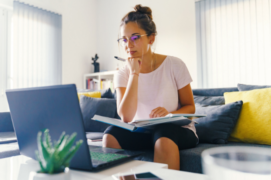 A woman wearing glasses sits on a couch, looking at a laptop with a pen in her mouth and an open notebook in her lap. She appears focused, surrounded by cushions and a small plant on the table.