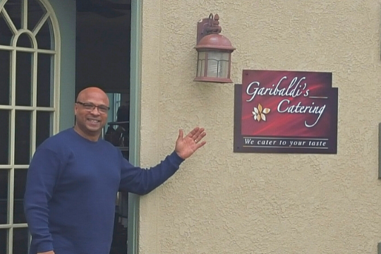 A smiling man in a blue sweater stands outside a building, gesturing towards a sign on the wall that reads Garibaldis Catering. We cater to your taste.