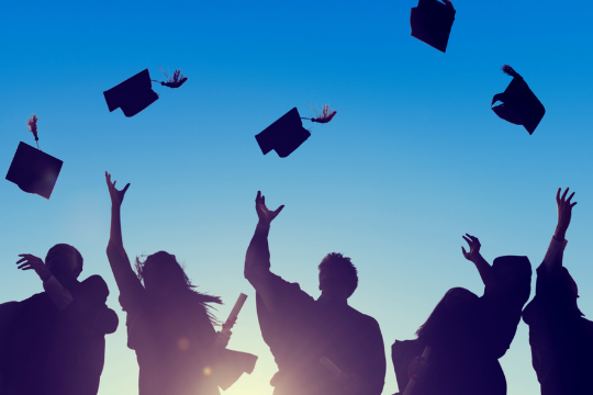 Silhouettes of graduates in gowns and caps celebrate by tossing their graduation caps into the air against a bright blue sky.
