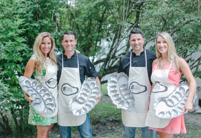 Four people, two men and two women, stand outdoors in front of green bushes, each wearing an apron and holding a large metal serving tray shaped for oysters or seafood. They are smiling at the camera.