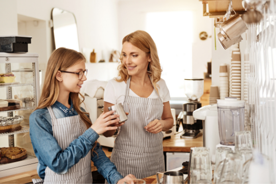 Two women wearing striped aprons stand in a bright café kitchen. One, holding a metal milk frother, smiles as they talk. Shelves, glassware, and baked goods are visible around them, creating a warm, inviting atmosphere.