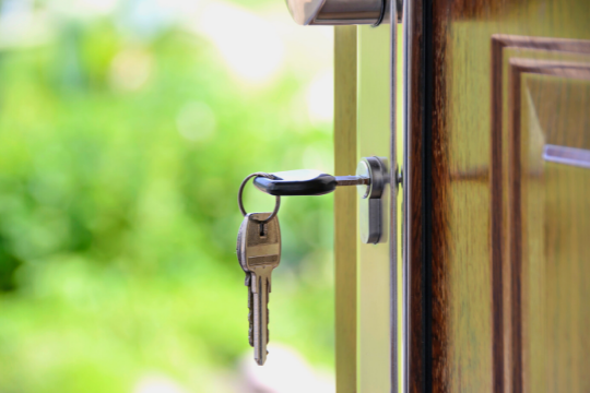 A close-up of a door that is slightly open with a key inserted in the lock; the key is attached to a keyring and two other keys. Greenery is visible outside in the blurred background.