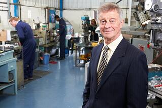 A man in a suit stands smiling in the foreground of a factory workshop, with workers operating machinery and assembling parts in the background.