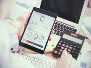 A person holds a tablet displaying graphs and charts in one hand while using a calculator with the other, with a laptop and documents visible on the desk.