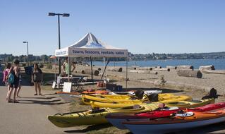 A group of people stand near a tent labeled “tours, lessons, rentals, sales” beside colorful kayaks on the ground by a beach, with water and driftwood logs in the background under a clear sky.