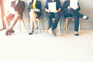 Four people in business attire sit on chairs in a row, holding resumes or documents, likely waiting for a job interview. Their faces are not visible, focusing on their lower bodies and attire.