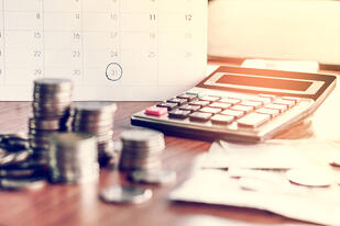 A close-up of stacked coins, a calculator, and scattered papers on a desk, with a calendar in the background marked on the 31st day of the month, suggesting financial planning or budgeting.