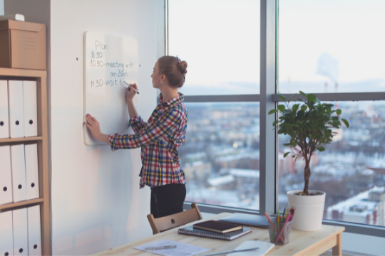 A woman stands in an office by a large window, writing on a whiteboard. There are shelves with binders, a desk with papers, and a potted plant nearby. A cityscape is visible outside the window.