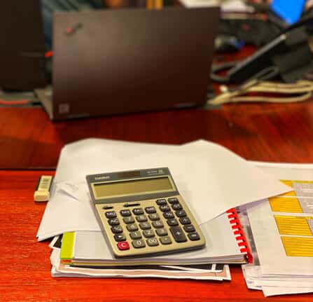 A calculator rests on top of scattered papers and a folder on a wooden desk, with a laptop, documents, and cables in the blurred background.