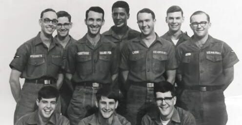 A black and white photo of ten smiling young men in military-style uniforms, standing and sitting in two rows, posing for a group portrait. The uniforms have name tags and patches on the chest and sleeves.