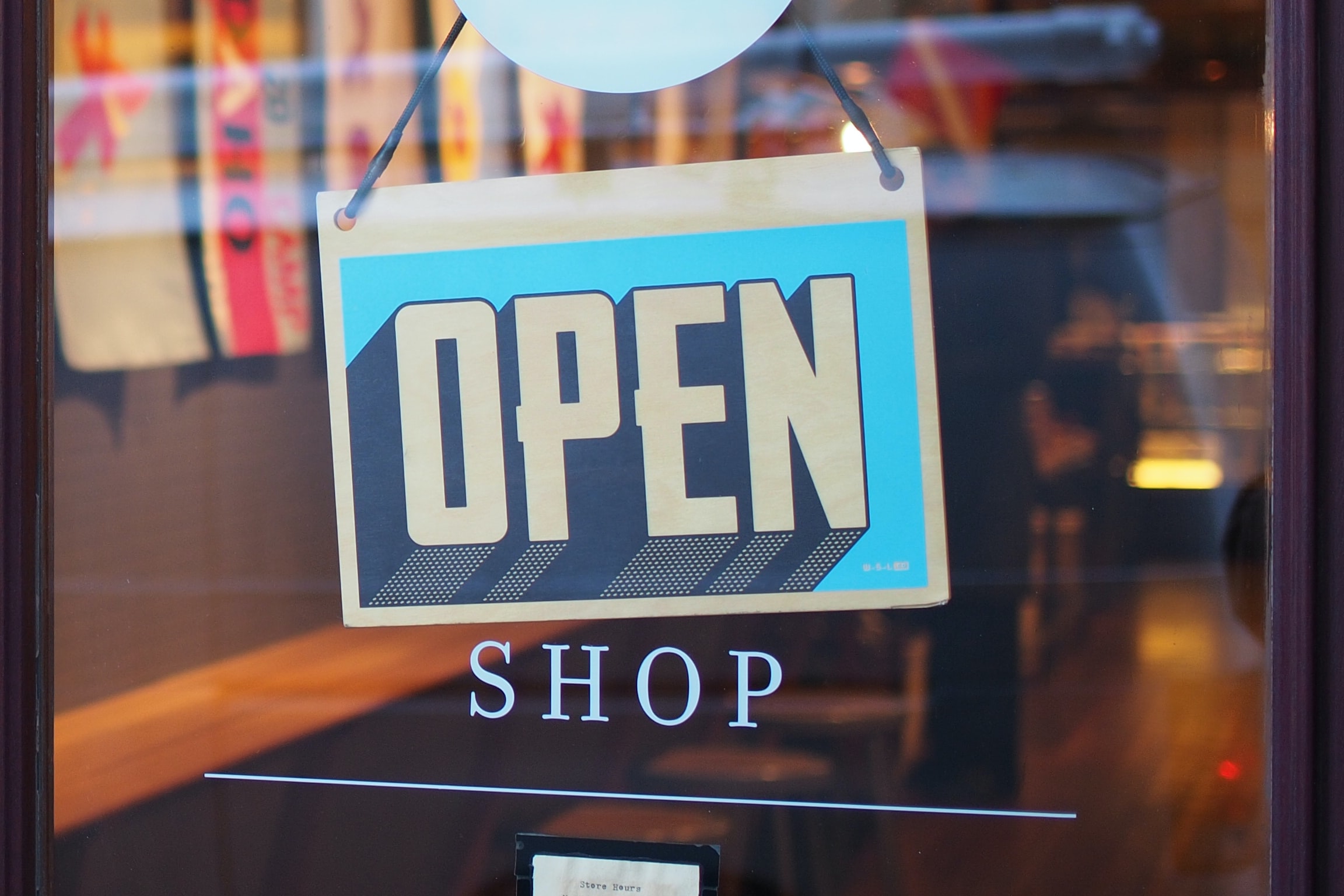 A blue and yellow OPEN sign hangs in the window of a shop, with the word SHOP in white letters on the glass below. The shop interior and colorful decorations are visible in the background.