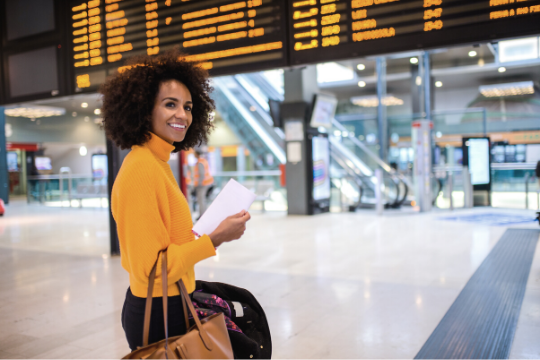 A woman in a bright yellow sweater stands in an airport terminal, holding travel documents and a bag, smiling while looking back, with a large electronic departures board visible above her.