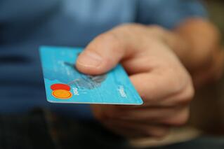 A close-up of a hand holding out a blue credit card, with the Mastercard logo visible in the corner, suggesting a payment or purchase transaction.