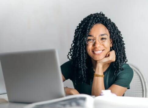A woman with curly hair and glasses smiles while sitting at a desk with an open laptop in front of her, resting her chin on her hand. A book and a water bottle are visible in the foreground.