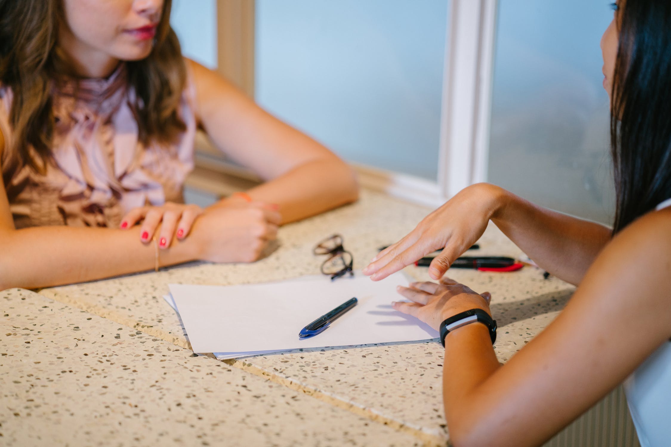 Two women sit across from each other at a table with paperwork, a pen, and glasses in front of them, engaged in conversation. Only their torsos and arms are visible.